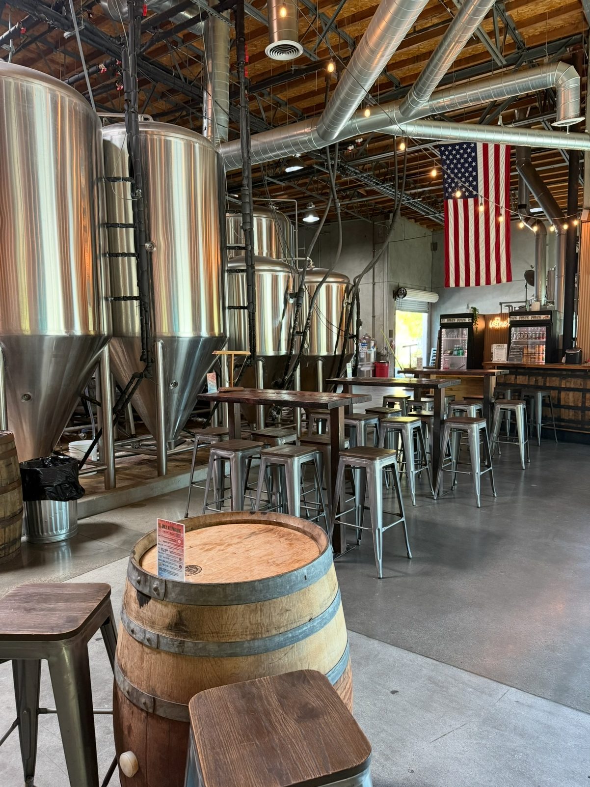The Prolific taproom floor beside the stainless fermenters, with barrel tables, high-tops and the bar
