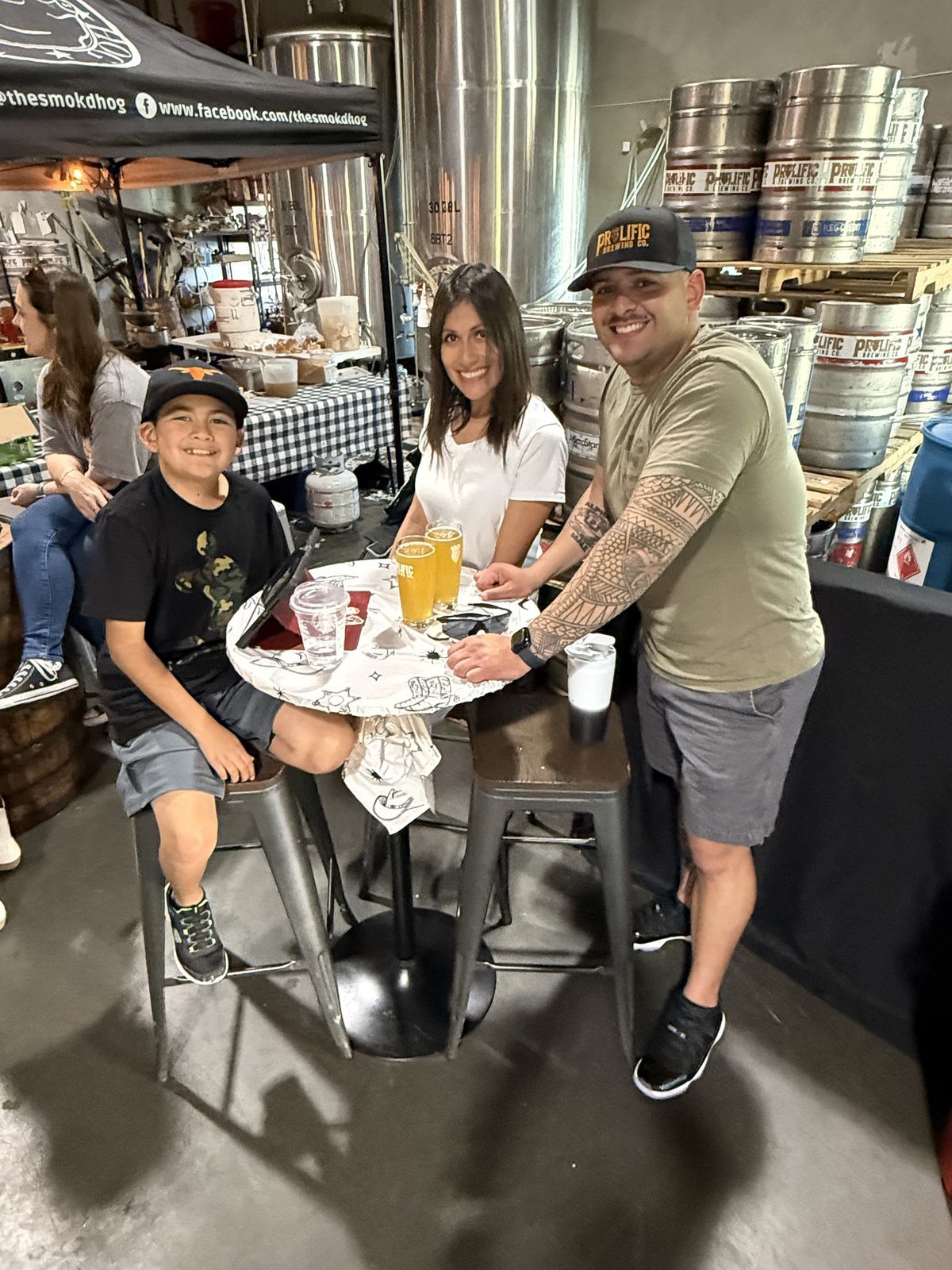 A family with their son at a high-top table in the brewhouse, kegs and fermenters behind them